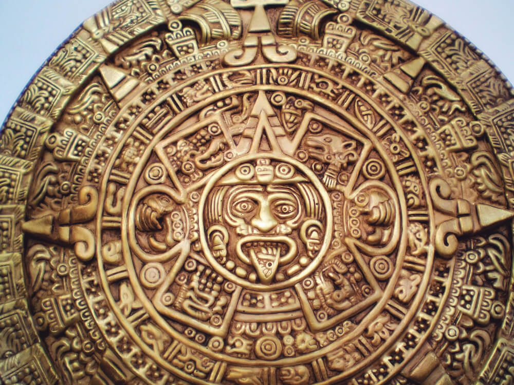 Fotografia. Calendário maia esculpido em pedra. Tom amarelado, formato circular, com diversas inscrições e relevos. Ao centro, um rosto com a boca aberta e a língua para fora.