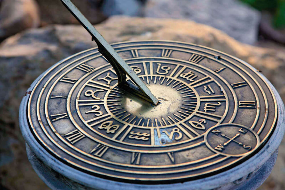 Fotografia. Relógio solar em metal envelhecido, estrutura circular e com marcações dos signos do zodíaco. O ponteiro fixado ao centro aponta para cima com ângulo de 20 graus.