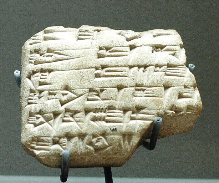 Fotografia. Tablete de pedra de formato irregular com escrita cuneiforme em suporte de metal preto.