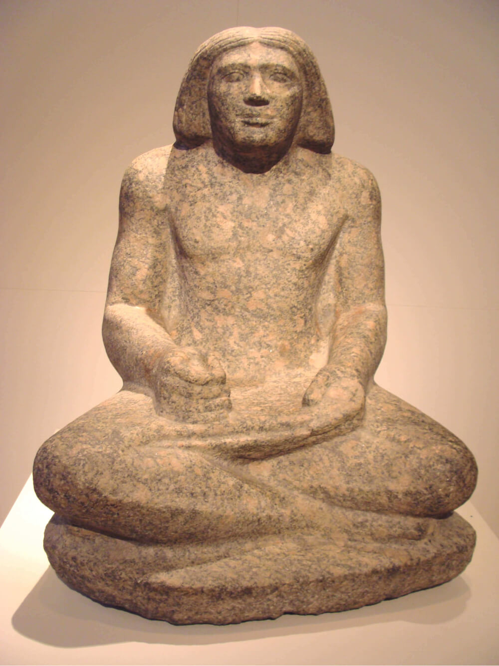 Fotografia de escultura em pedra. Homem escriba sentado de pernas cruzadas. Cabelo liso, na altura dos ombros. Braços apoiados nas pernas. Mãos seguram um papiro.